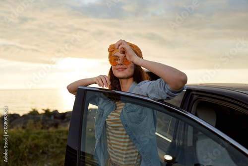 Young woman leans from a car window at sunset, holds a round pastry to her face, wearing a striped tee and denim jacket, radiating a carefree road trip mood.