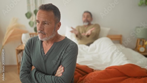 Man with arms crossed sits beside bed in building while a second man gestures from under covers and speaks, looking away; relationship tension.