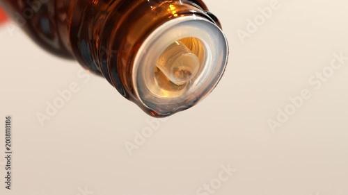 A drop of essential oil slowly moves from a brown bottle, beige background.