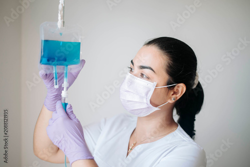 Female nurse wearing protective gloves and face mask carefully inspecting an IV fluid bag before patient treatment in a modern healthcare environment.
