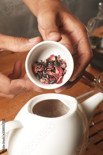 Slika na platnu Hands holding a small ceramic bowl filled with loose-leaf herbal tea containing dried rose petals, preparing to brew it in a white teapot