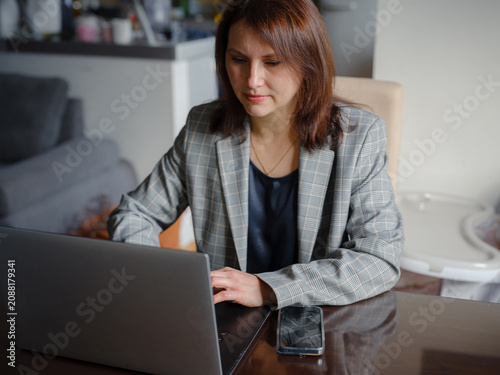Mother working remotely from home, multitasking between professional tasks and family responsibilities, using a laptop in a cozy home environment.
