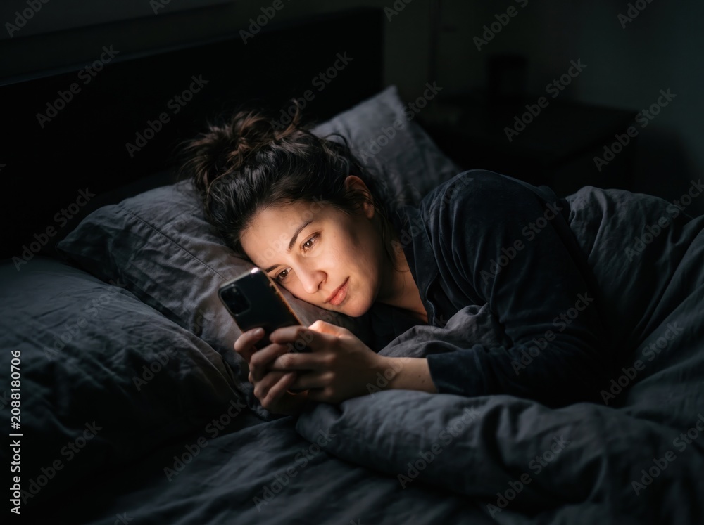 Fototapeta premium Scrolling through the Night: A woman lies in bed, illuminated by the dim light of her smartphone, engrossed in its screen as darkness envelops the room.