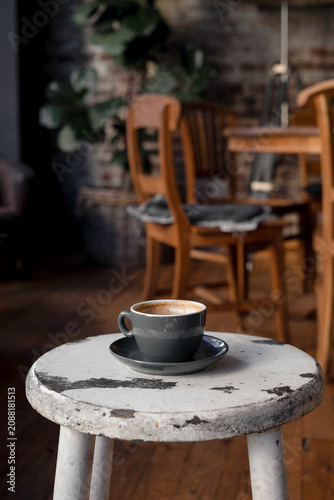 Cup of cappuccino on a weathered white wooden stool in a cozy café interior. Warm natural light, shallow depth of field, visible film grain and analog-inspired aesthetic create a calm.