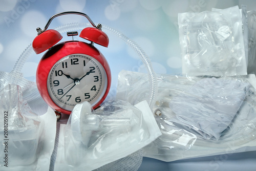 A red alarm clock sits on a table surrounded by silicone male catheters and urine bags.