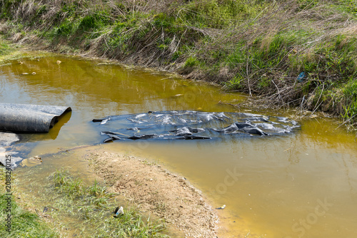 A small creek shows signs of pollution with dark material floating on the water. The area has grass and shrubs along the banks
