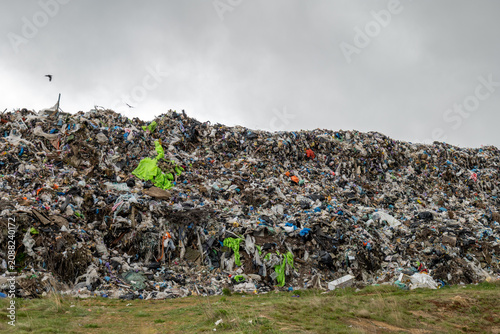 Piles of waste rise at a landfill. Workers in bright clothes can be seen sorting through the trash on a cloudy day