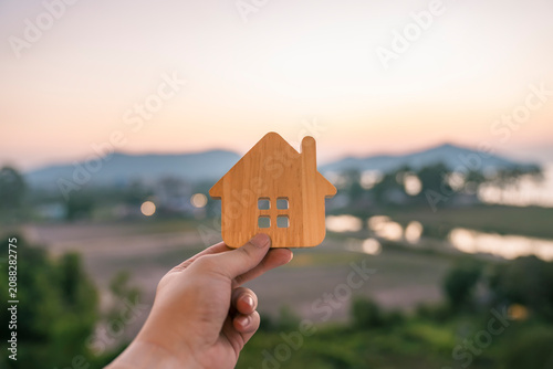 Hand holding a small wooden house icon with sunset nature background. Real estate property, insurance, home mortgage loan, refinance, eco living and family land concept.