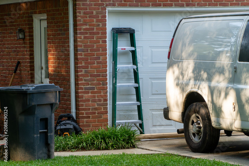 Canvastavla White utility van parked in a sunny residential driveway with a green stepladder leaning against a white garage door, next to a brick house, trash can, and lawn equipment