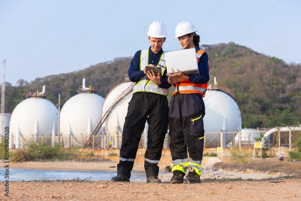 Obraz premium Two engineers safety helmet and reflective vest holding laptop and tablet standing near large spherical storage tank industrial facility with focused expression discussing project details outdoors