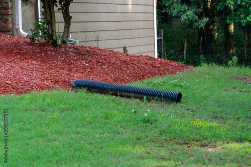 Fotografía Black corrugated drainage pipe on lush green lawn leading from a house downspout, bordered by vibrant red wood chip mulch and a residential home exterior
