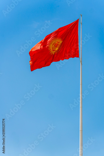 The national flag of the Kyrgyz Republic waving on a flagpole against blue sky. Low angle view of Kyrgyz red flag with a yellow sun and tunduk on a high flagpole.