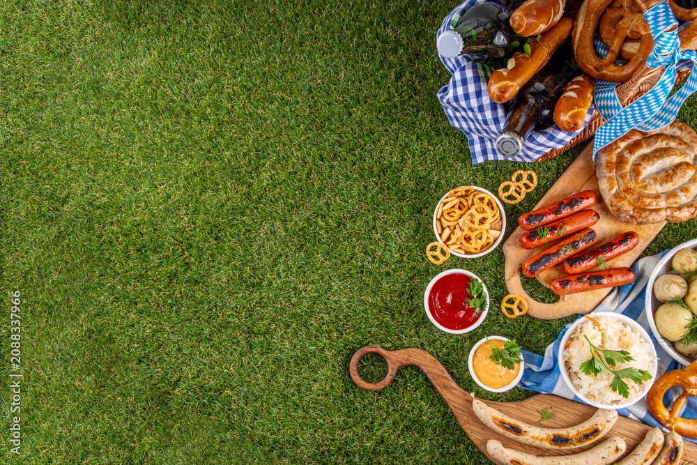 Fototapeta premium Oktoberfest picnic and barbecue food, bavarian party menu, bavarian sausages, pretzels, potato, sauerkraut, beer bottles, with picnic basket, checkered napkin tablecloth, Oktoberfest festive ribbon