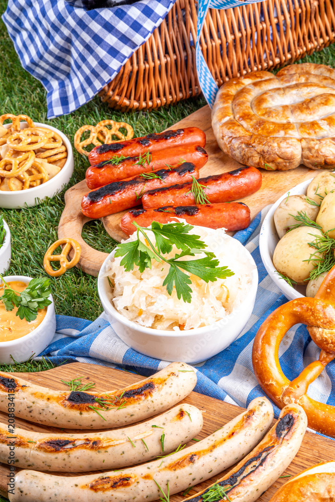 Fototapeta premium Oktoberfest picnic and barbecue food, bavarian party menu, bavarian sausages, pretzels, potato, sauerkraut, beer bottles, with picnic basket, checkered napkin tablecloth, Oktoberfest festive ribbon