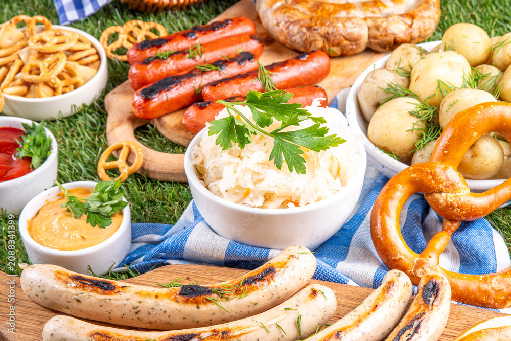 Fototapeta premium Oktoberfest picnic and barbecue food, bavarian party menu, bavarian sausages, pretzels, potato, sauerkraut, beer bottles, with picnic basket, checkered napkin tablecloth, Oktoberfest festive ribbon