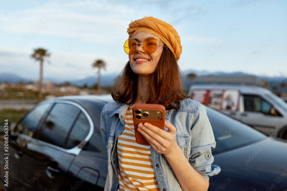 Naklejka premium A woman lifestyle casual travel sunny phone sunglasses turban denim outdoor scene with a cheerful pose near parked cars in bright daylight.