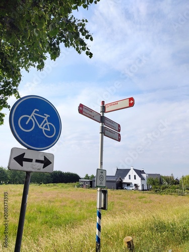 A red bicycle Dutch direction sign, right going to Noordwijk, Sassenheim and Den Haag, left to Centrum and Haarlem, and middle pointing to the beach. Next to it a traffic sign for cycling lane. 