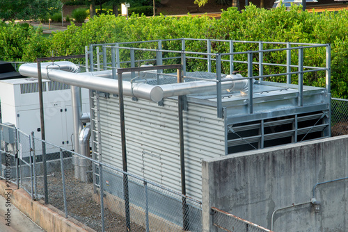 Фотография Large outdoor industrial cooling system with insulated pipes, a generator, and metal railings, enclosed by a chain-link fence amidst green foliage for commercial HVAC infrastructure