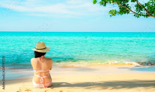 Bild auf Leinwand Woman in a pink bikini sitting on tropical beach facing turquoise ocean beneath a shady tree
