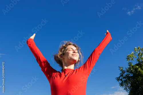happy, free-spirited young European woman (aged 18 25) wearing a red long-sleeved top enjoys moment, standing in sunlight under cloudless sky. She loves life, dreaming, values positive mindset