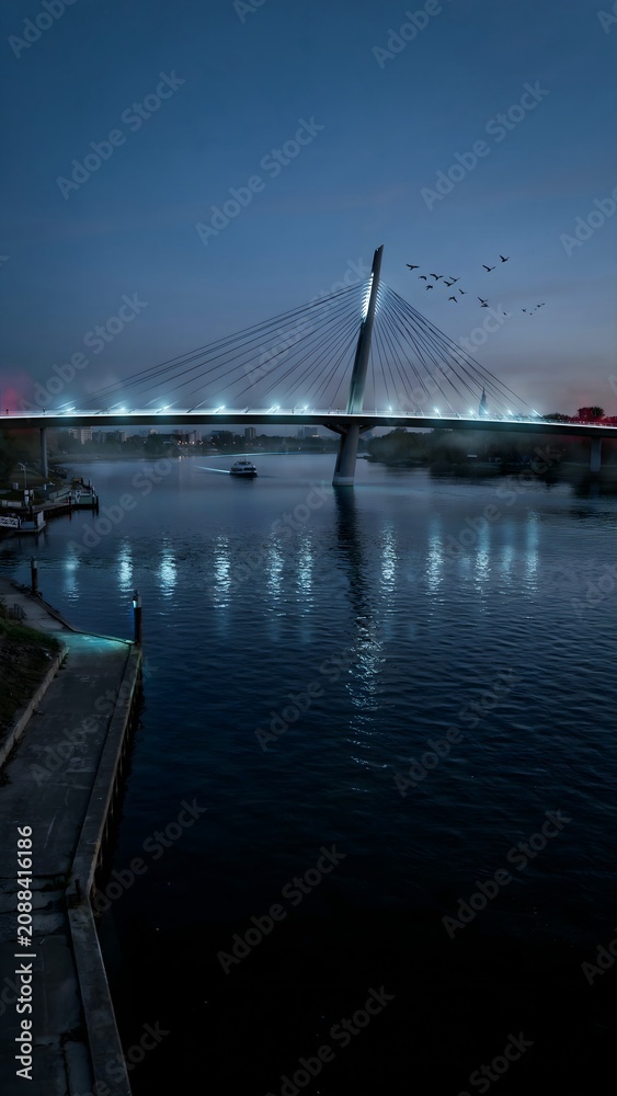 Fototapeta premium Modern cable-stayed bridge over river at dusk with city skyline and boats