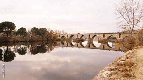 Masonry Pointed arch Stone bridge (Puente de Piedra) across the river Duero in Zamora, Castile-Leon, Spain. Reflection in the water. Autumn nature. Pedestrians walking on the bridge. Overcast.
