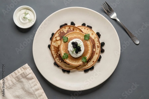 Plate of Blini with Caviar garnished with sour cream and blackberries. Concept of thin pancakes with caviar sour cream on gray surface with linen napkin and fork.
