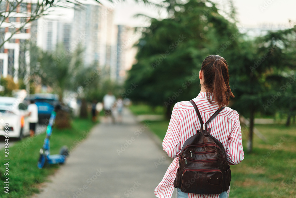 Naklejka premium Woman lifestyle, walking away with backpack along a sunlit city path, casual stride, greenery beside the route, candid moment, relaxed mood, natural colors