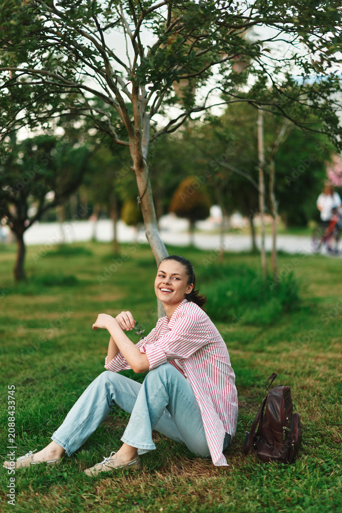 Naklejka premium A smiling young woman student sits on the grass in a park, wearing casual clothes with a backpack nearby. Concept of student life and outdoor leisure.