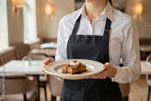 Waitress in white shirt, black apron holding Sticky Toffee Pudding topped with cream. Concept of cozy restaurant atmosphere, date sponge with toffee sauce cream in focus.