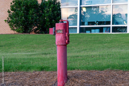 Tableau sur toile Vibrant red fire standpipe with 'OPEN' sign, set on green grass with pine straw mulch, against a modern building with reflective windows and lush green landscaping