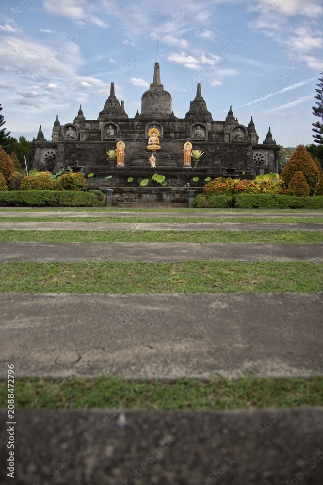 Obraz premium Magnificent mini replica of Borobudur temple surrounded by lush gardens, pathways, and golden Buddha statues under a blue sky at Brahma Vihara Arama monastery in Bali.
