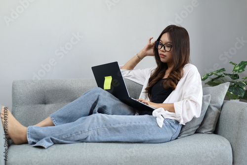 фотографія A young Asian woman sits on the sofa in her living room, working on her laptop while holding her head with a tired, lethargic and stressed expression due to a work deadline