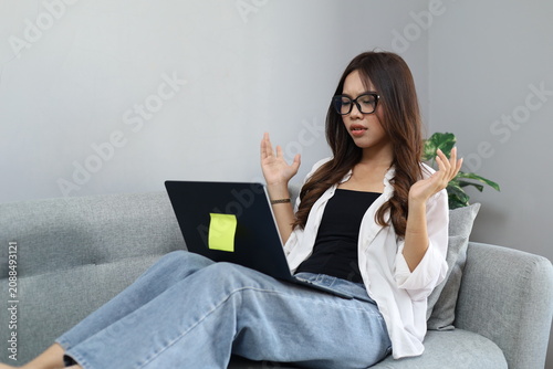 Foto A young woman sits on a sofa in the living room while working, staring anxiously at a laptop screen, reacting to bad news, feeling frustrated and angry, while shrugging her shoulders and hands