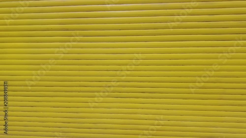 The camera moves backward from a bright yellow metal roller shutter door. The physical movement gradually reveals the structured horizontal texture of the protective security blinds.