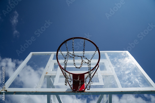 Outdoor basketball hoop with metal chain net against bright blue sky and clouds, low angle view highlighting urban court detail and sporty recreation, with basketball, hoop, rim, chain detail
