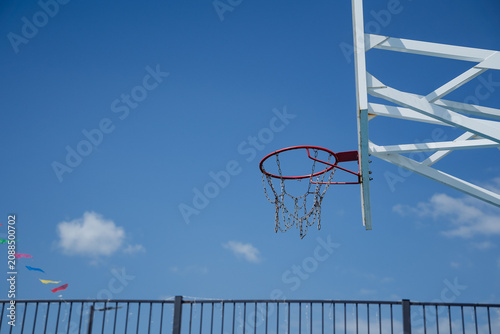 Outdoor basketball hoop with red metal rim, chain net, white support beams, and clear blue sky background viewed from below, with basketball, rim, chain, net detail