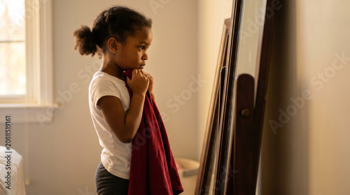 Young black girl holding red cloth while looking in mirror at home  