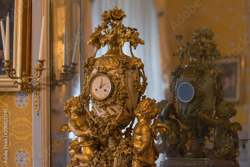 An ornate, gilded mantle clock featuring cherub figures stands as a sophisticated centerpiece, its intricate craftsmanship reflected in a nearby mirror to create a sense of timeless elegance and luxur