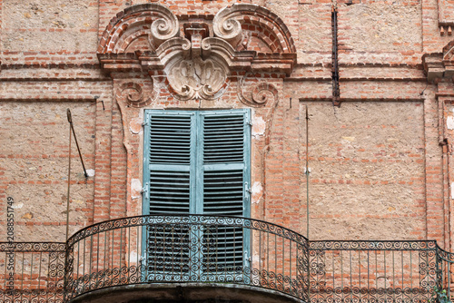 A detailed view of a rustic red brick building facade, highlighting a classical window adorned with an ornate pediment and a wrought-iron balcony railing