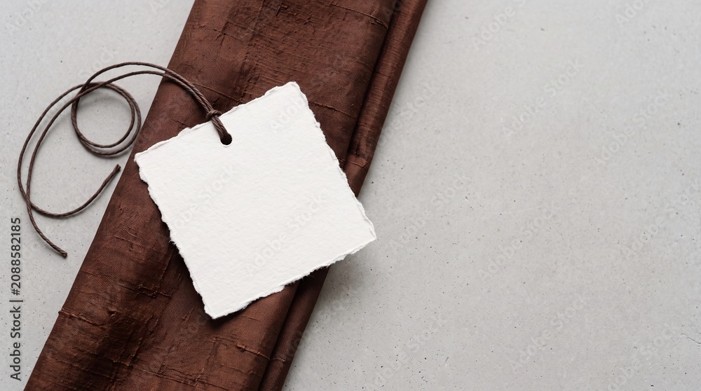 Fototapeta premium A blank white tag with a rough edge is tied with a brown string. The tag rests on a brown fabric surface, creating a simple and elegant composition.