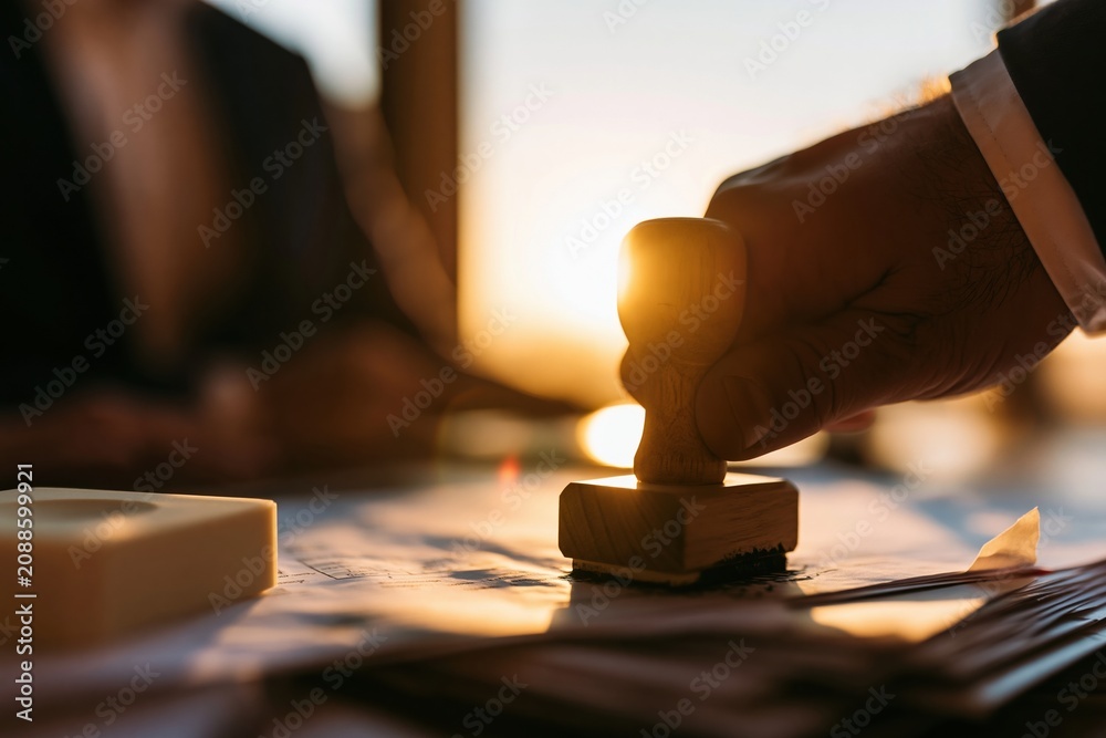 Obraz premium Close up of a hand pressing a wooden stamp onto official paperwork for a visa application, with warm golden sunlight creating a glowing halo for immigration and legal services