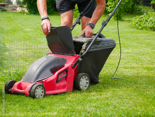 Man gardener operating a red electric lawn mower on a green grass lawn. Maintenance of backyard garden territory. Concept of landscaping and summer lawn care work in spring time.