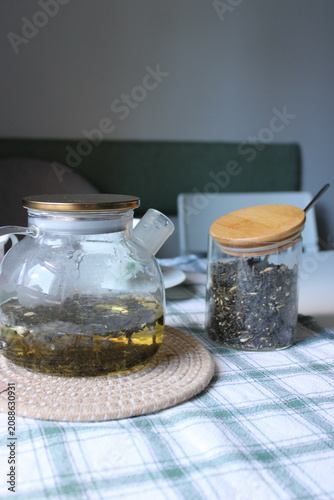 Glass teapot filled with brewed herbal tea beside a jar of loose tea leaves on a checkered tablecloth in a cozy indoor setting