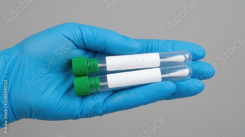Hand in blue latex glove holding two test tubes with green caps and cotton swabs, suitable for medical testing or laboratory analysis of viruses like hantavirus and ebola