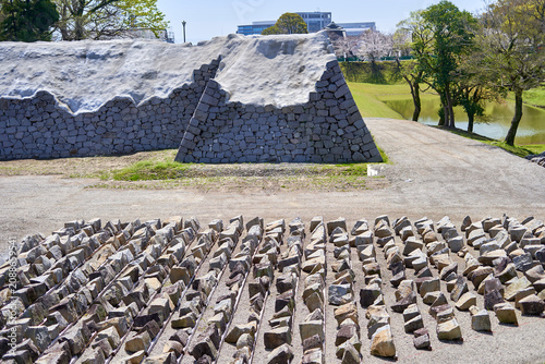 Stone walls of a historic Japanese castle undergoing restoration, with numerous cataloged stones arranged in organized rows on gravel in the foreground. The ancient stone fortification features a