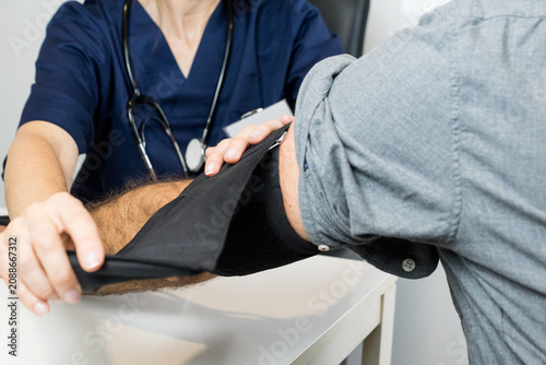 Obraz na plátně Female physician in scrubs tightening the strap of a manual blood pressure monitor on a male patient sitting at a clinical desk