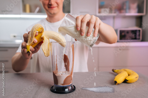 Young man creating healthy banana smoothie, peeling banana into blender while accidentally spilling milk from glass, causing small mess on kitchen counter.