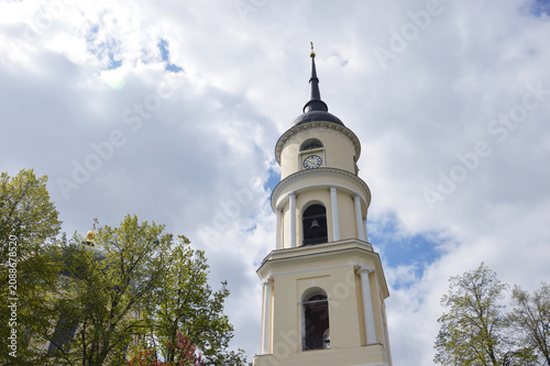 Bottom view of a yellow orthodox bell tower with clock and spire against cloudy sky.