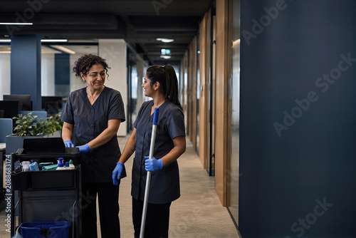 Two cleaners standing in office hallway. Women staff with cleaning trolley and mop equipment talking. Professional janitorial service and maintenance team working in modern workspace.